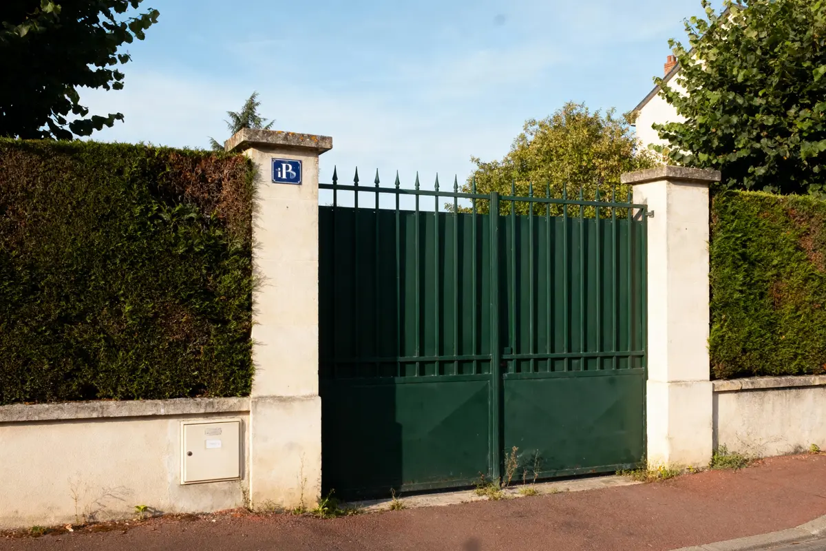 Portail vert en fer, entouré de murs blancs et haies, sous un ciel bleu.