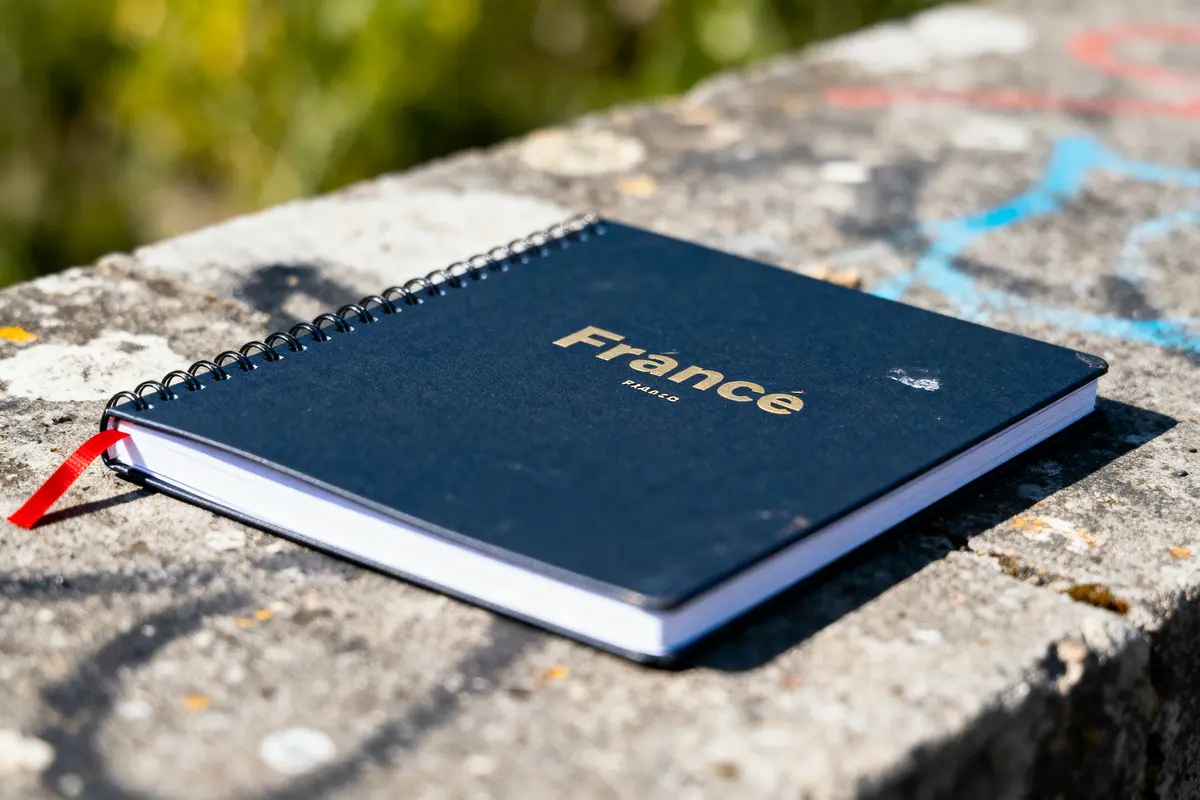 Carnet bleu avec inscription "France" sur un mur en béton grafité, éclairé par le soleil.