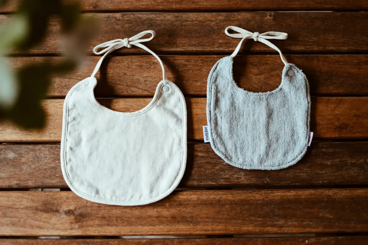 Deux bavoirs en tissu, un blanc, un gris, posés sur une table en bois brun.