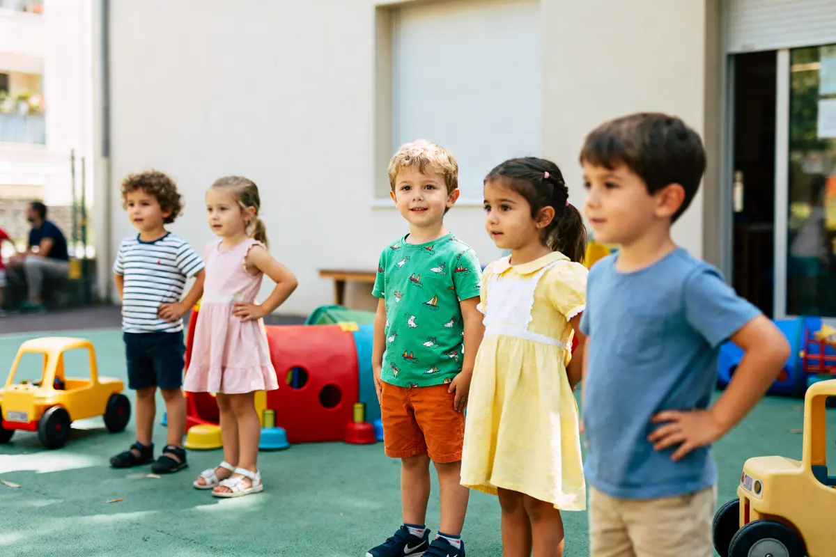 Cinq enfants debout dans une cour de récréation avec des jouets en arrière-plan.