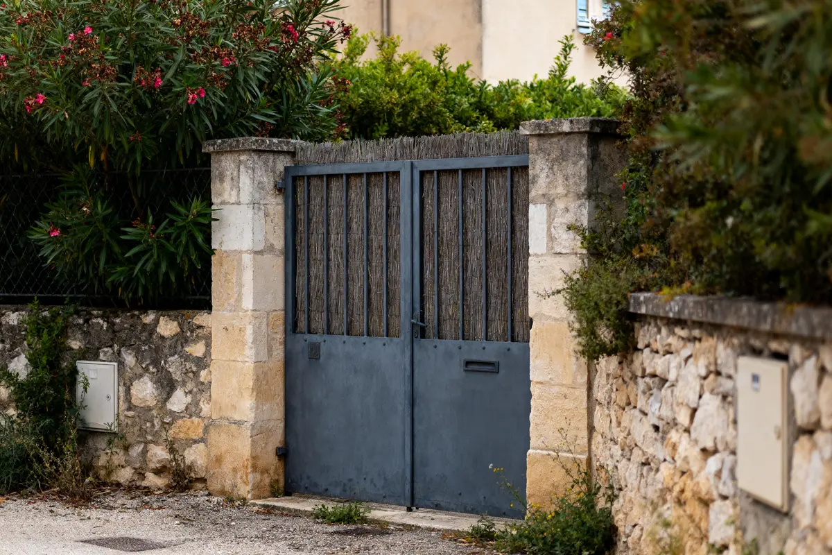 Portail métallique bleu entouré de murs en pierre et végétation verdoyante avec fleurs roses.