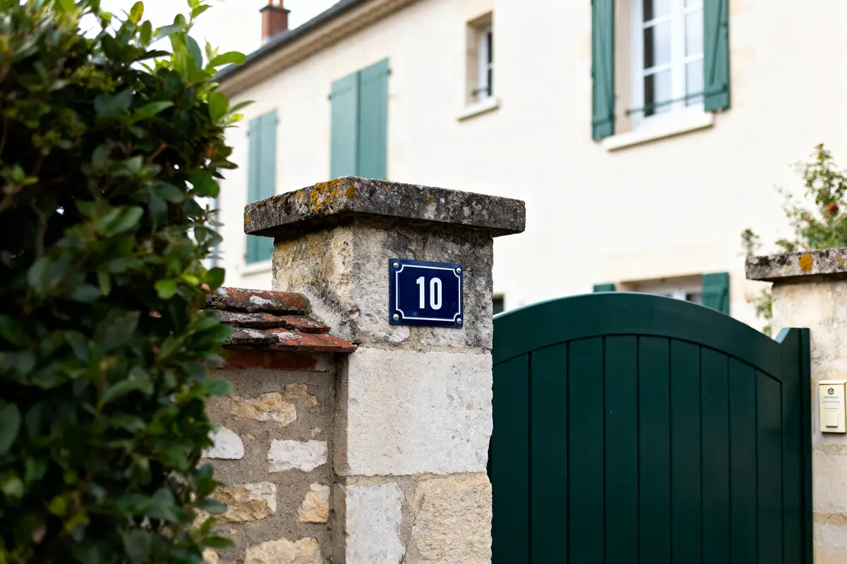 Mur en pierre avec numéro 10, portail vert, volets verts, maison en arrière-plan.