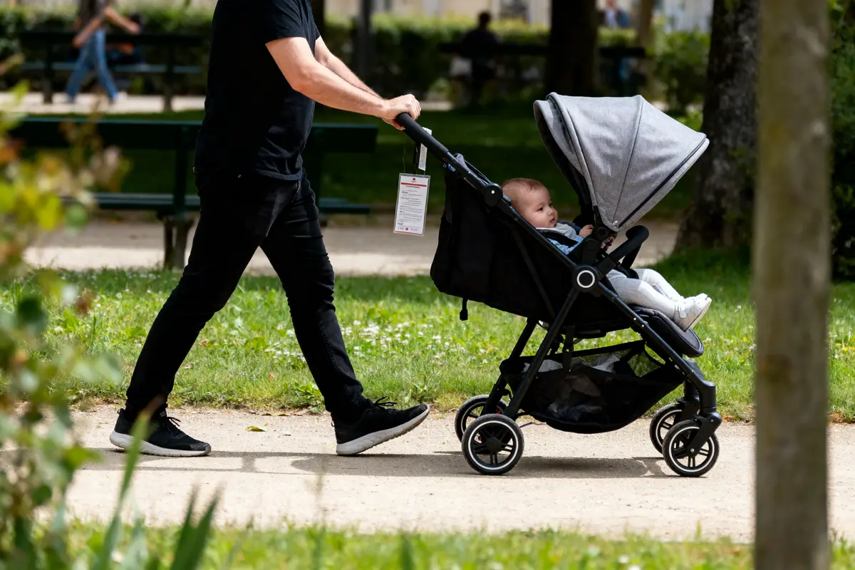 Personne en noir poussant une poussette avec bébé, dans un parc vert et ensoleillé.